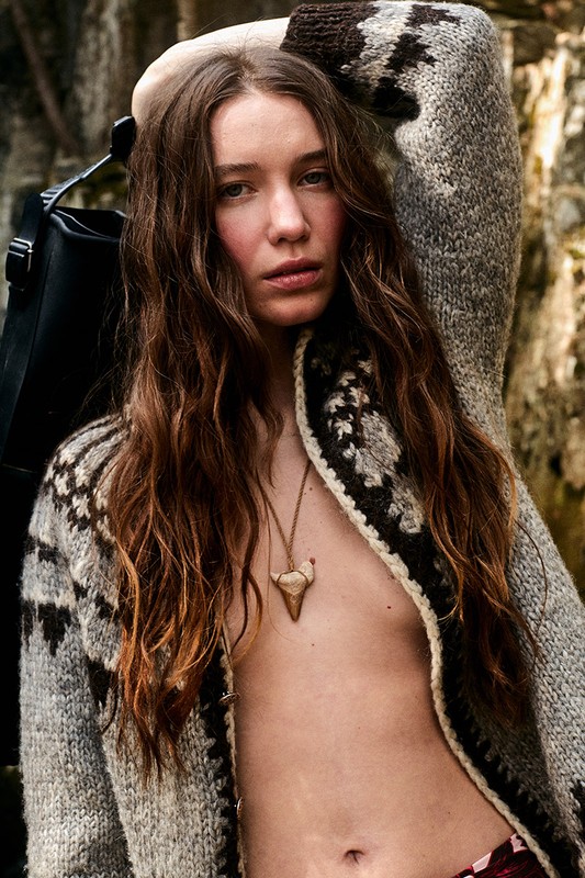 Woman with long brown hair poses in a patterned gray cardigan, wearing a tooth necklace, against a muted, natural backdrop.