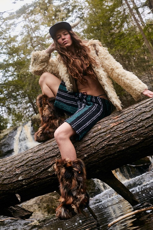 A woman with long hair poses while sitting on a fallen tree wearing fur boots, shorts, and a shaggy jacket with a forest background.