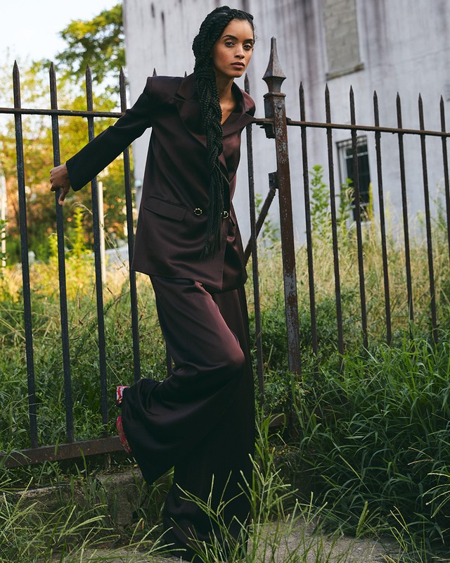Woman with braids poses in a brown suit and pink shoes outdoors in front of a rusted fence, overgrown grass, and a building.