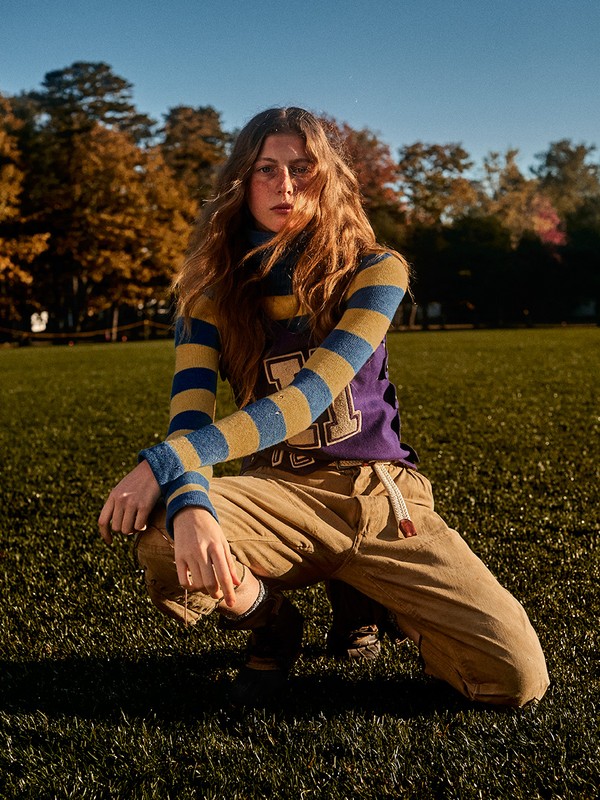 A woman with long brown hair poses in a grassy field wearing a striped shirt, sweater vest, and tan pants, illuminated by natural light.