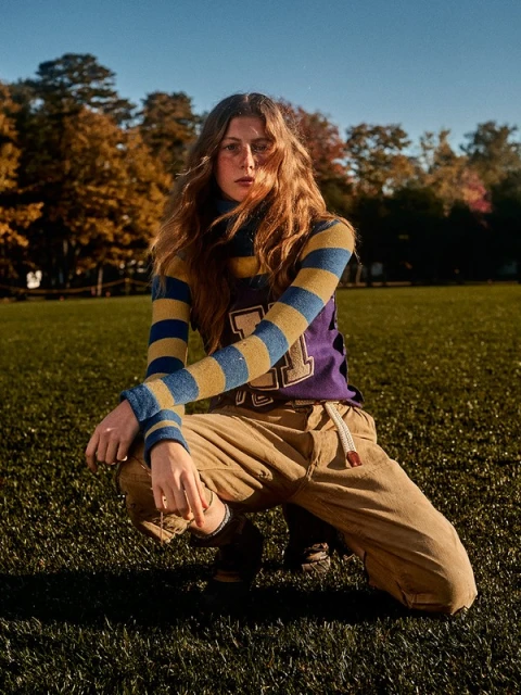 A woman with long brown hair poses in a grassy field wearing a striped shirt, sweater vest, and tan pants, illuminated by natural light.