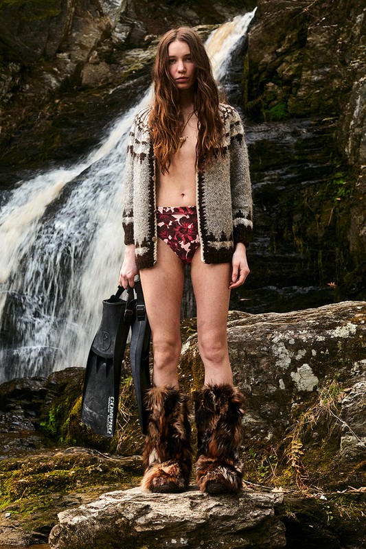 Woman in swimsuit, fur boots, and cardigan stands near waterfall, holding flippers, in outdoor fashion shot with natural lighting.