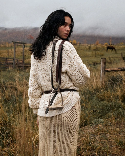 A woman in a cream sweater and skirt stands in a grassy field, glancing over her shoulder with a horse visible in the background.