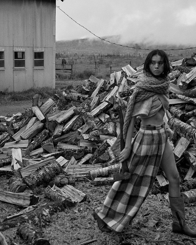 A woman in a skirt and shawl poses near a pile of logs in a black-and-white, slightly overcast rural outdoor setting.