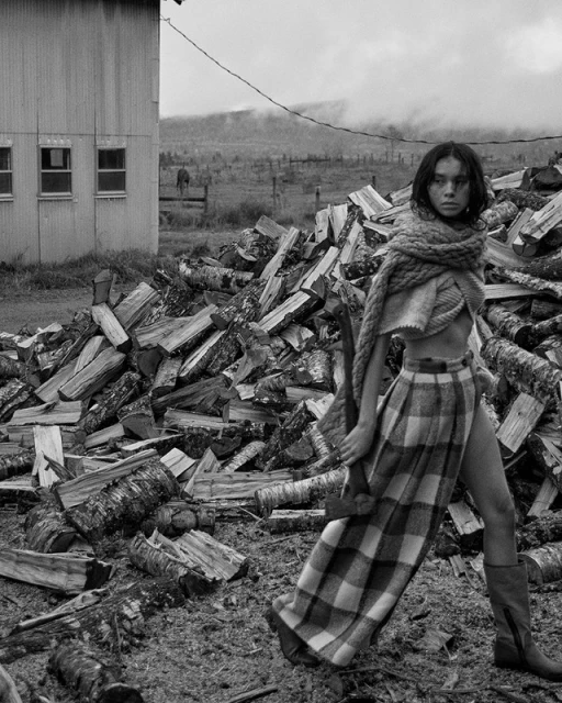 A woman in a skirt and shawl poses near a pile of logs in a black-and-white, slightly overcast rural outdoor setting.