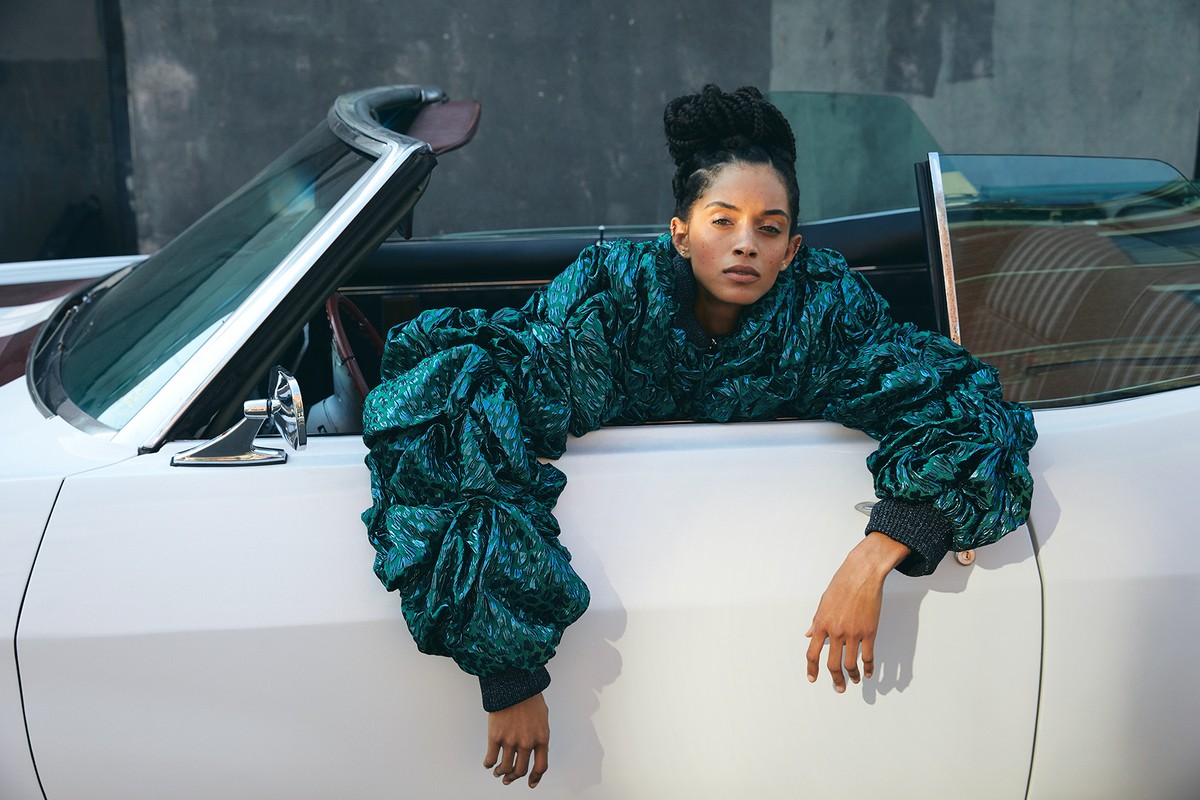 Fashionable adult female poses in an open convertible, wearing a shiny green jacket with large sleeves, in an urban outdoor setting.