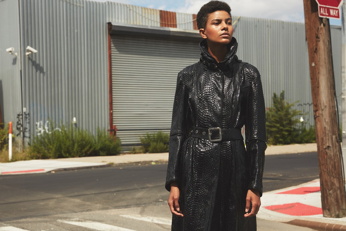 An adult female with short hair poses on a city street wearing a black snake-skin-pattern coat with a black belt and silver buckle.