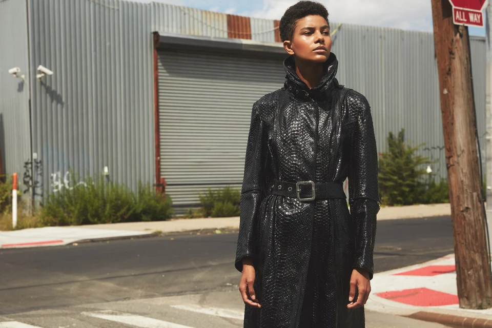 An adult female with short hair poses on a city street wearing a black snake-skin-pattern coat with a black belt and silver buckle.