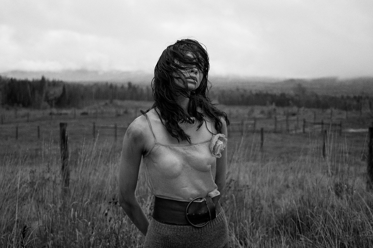 Monochrome portrait of a woman with dark hair in a spaghetti-strap top and wide belt, standing in a field with trees and sky in the background.