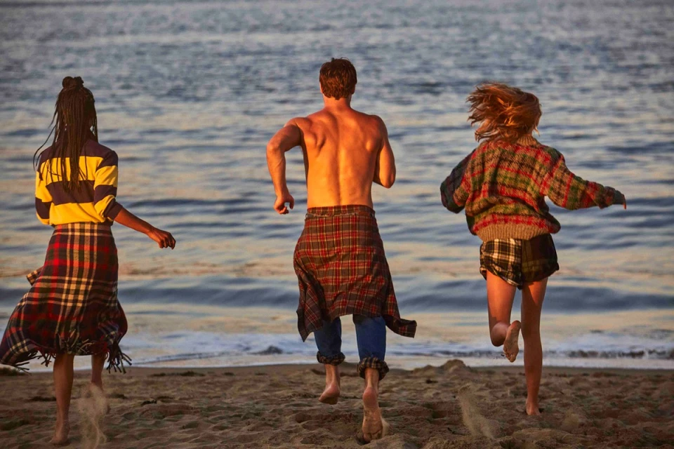 Three young people run barefoot on a sandy beach toward the ocean at sunset, wearing casual plaid clothing, enjoying a beach day.