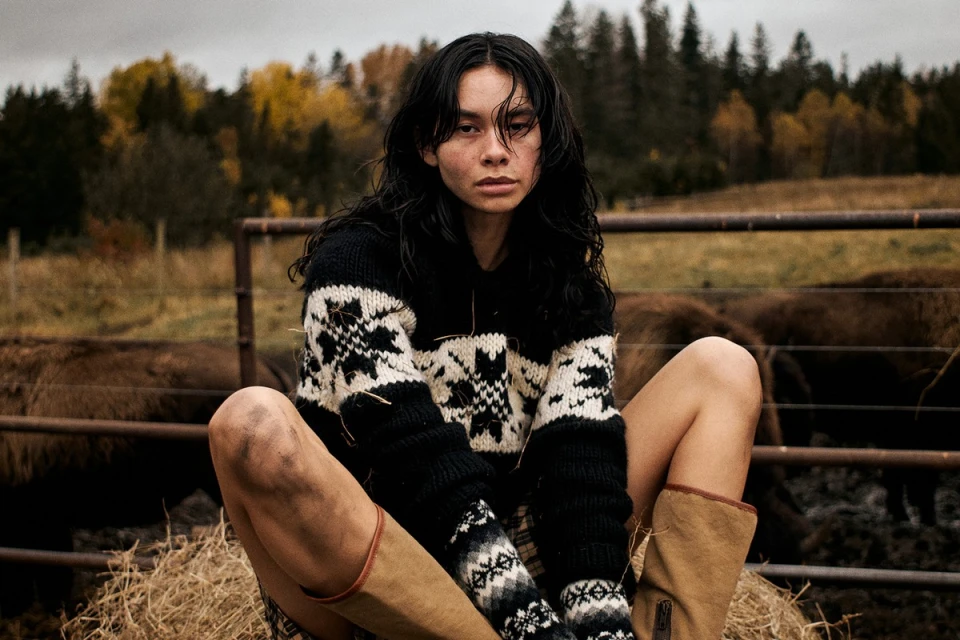 Woman with long dark hair poses in a patterned sweater and boots on hay, in a rural setting with cows and autumn trees.