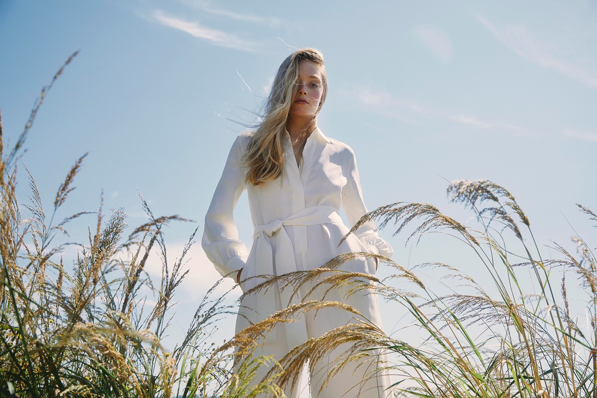 A young woman in a white jumpsuit stands in a field of wheat-colored tallgrass against a sunny blue sky in a fashion editorial shot.