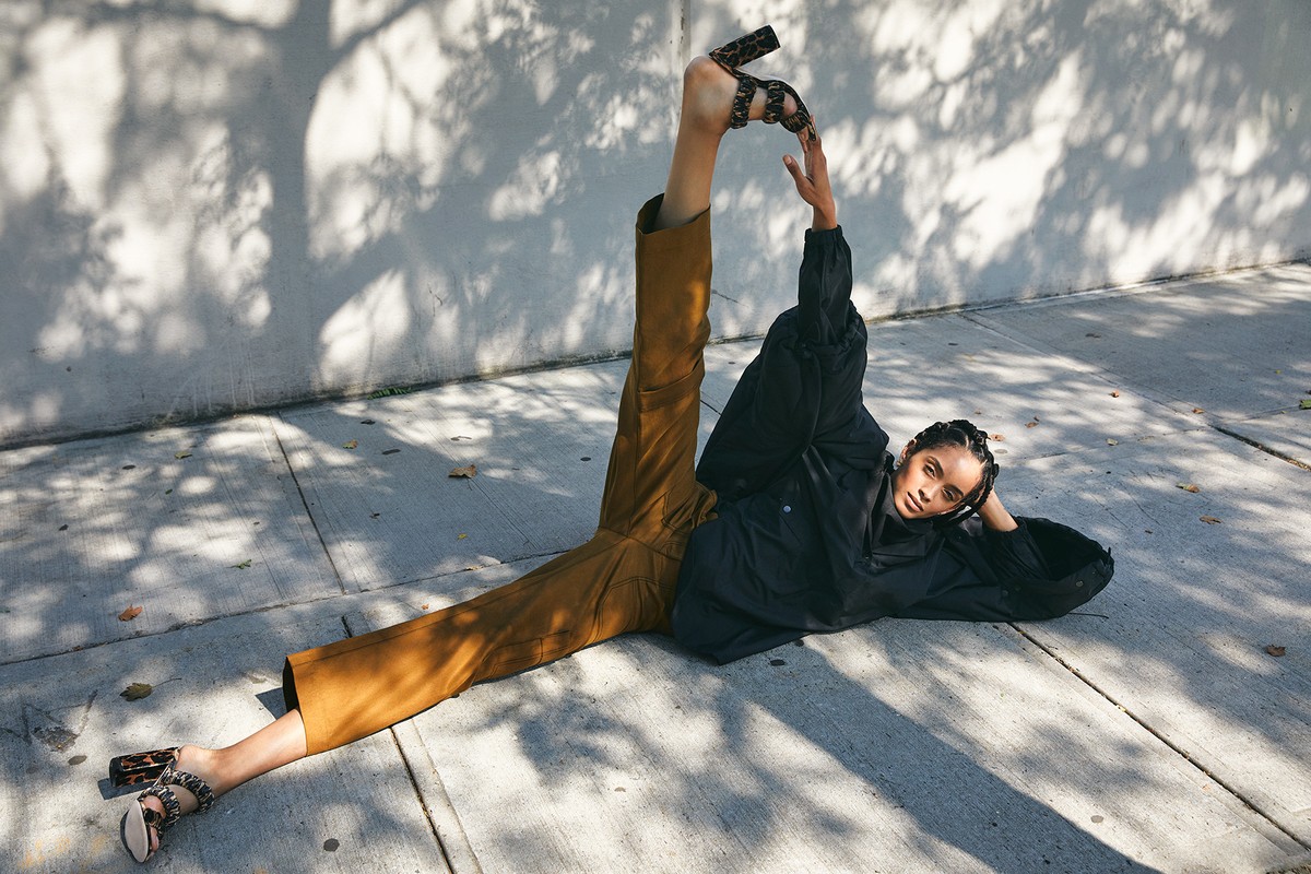 Woman in black top and tan pants poses creatively on concrete with tree shadows, wearing heeled sandals, showcasing fashion and style.