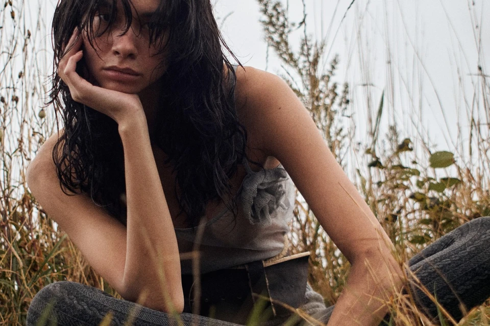 A young adult woman with dark, wet hair and a neutral expression sits outside surrounded by tall, golden grasses in a medium shot.