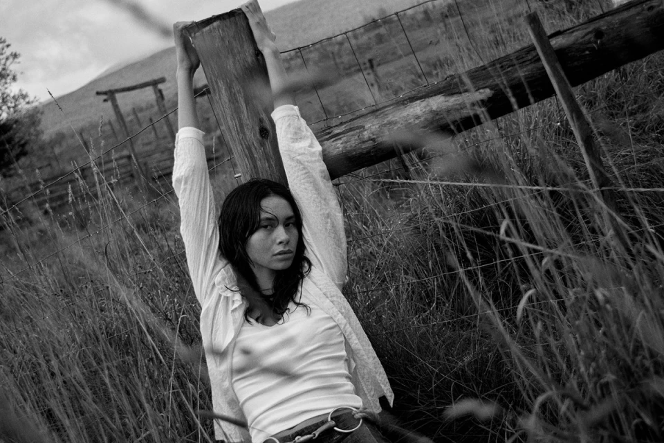 Monochrome portrait of a young woman with long dark hair in a white blouse, holding onto a wooden fence post surrounded by tall grass.