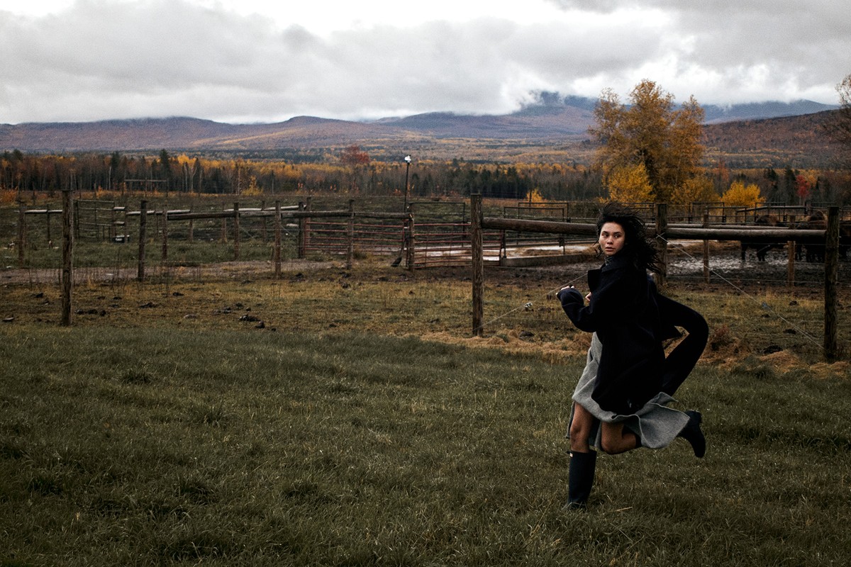 A woman runs across a grassy field with mountains and trees in the background. She wears a coat, skirt, and boots.