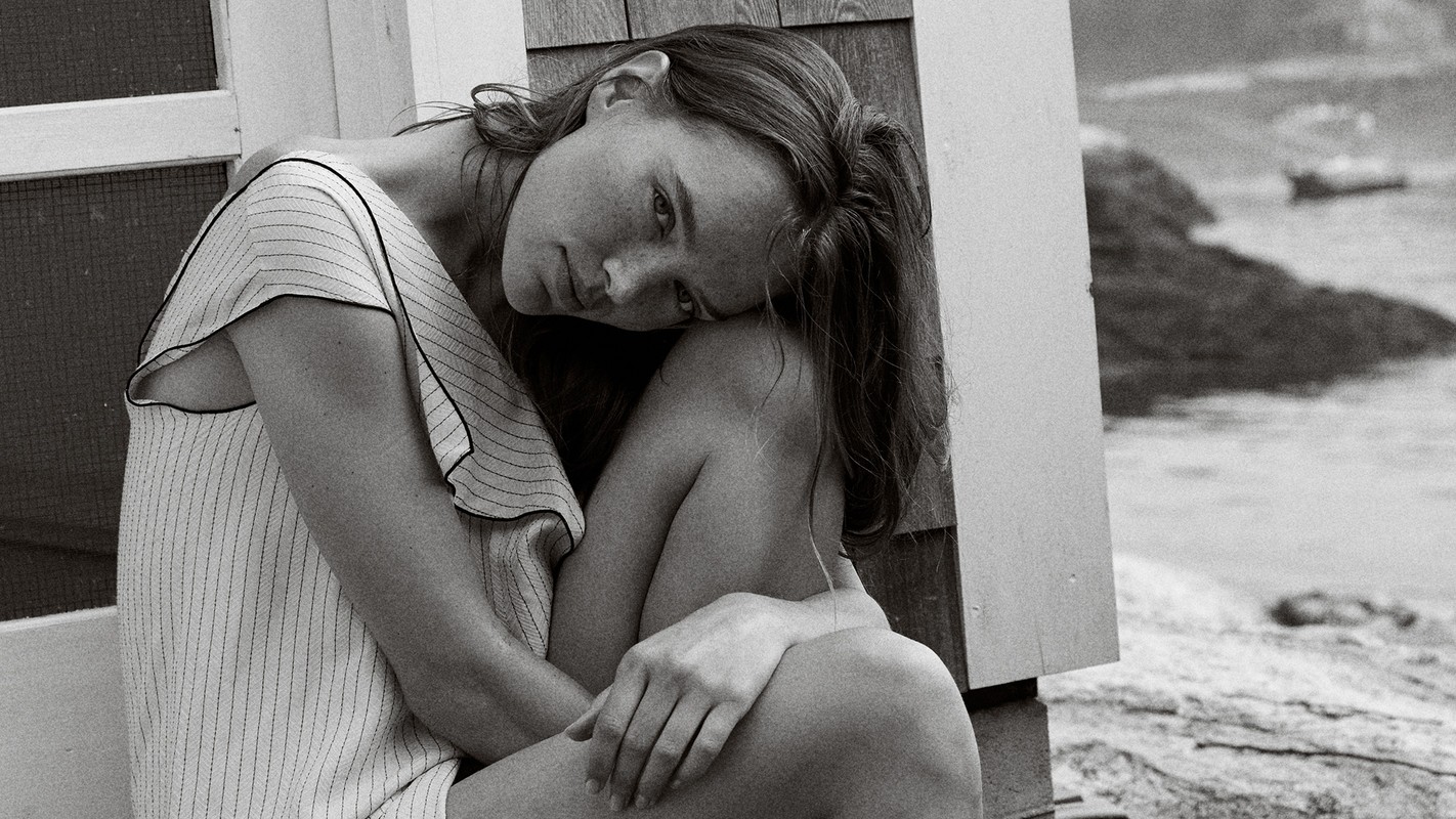 Black and white portrait of a woman with her head resting on her knees sitting outside near water, creating a serene and contemplative scene.
