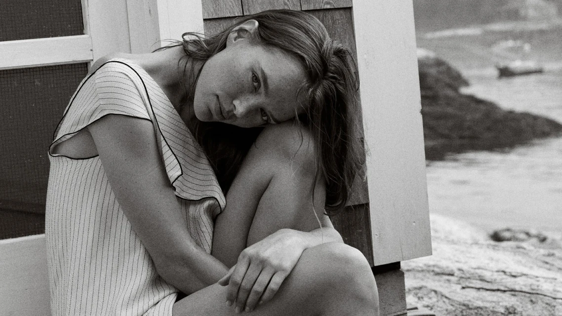 Black and white portrait of a woman with her head resting on her knees sitting outside near water, creating a serene and contemplative scene.