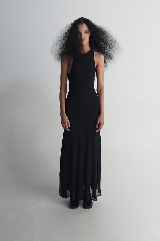 Woman with dark curly hair poses in a full-length black halter dress in a studio with a white background, looking towards the camera.