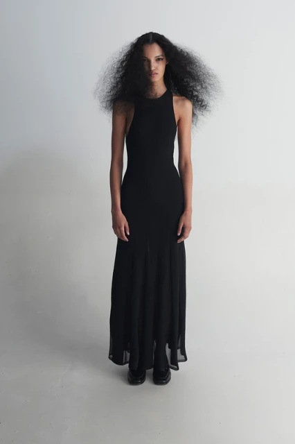 Woman with dark curly hair poses in a full-length black halter dress in a studio with a white background, looking towards the camera.