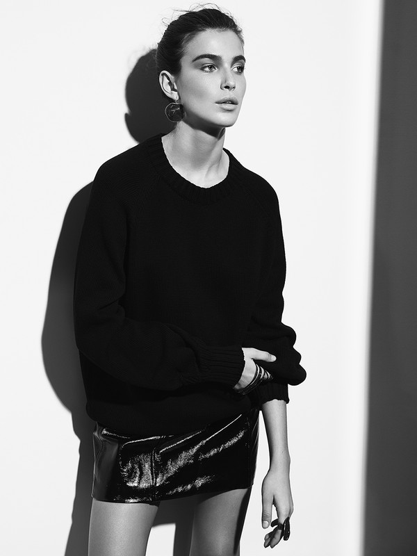 Black and white portrait of a woman wearing a dark crewneck sweater and shiny miniskirt against a wall.
