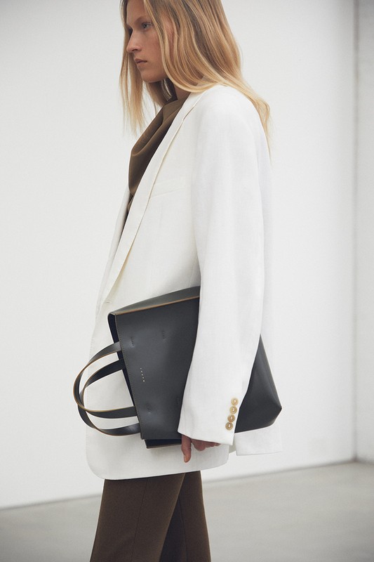 Woman in a white blazer holding a black leather tote bag, posing in front of a white wall. Fashion editorial photography.