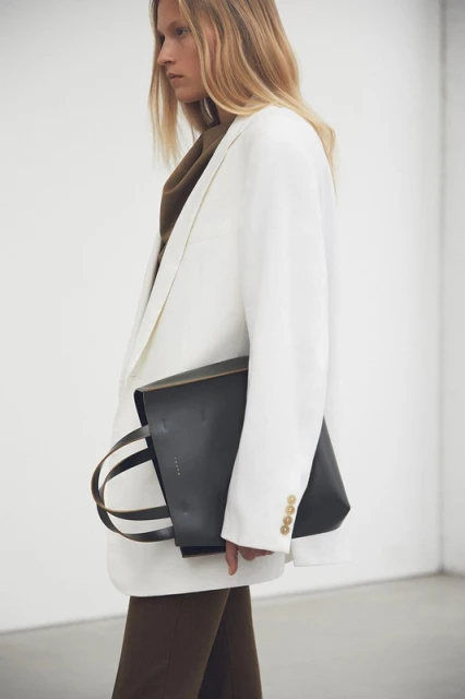 Woman in a white blazer holding a black leather tote bag, posing in front of a white wall. Fashion editorial photography.