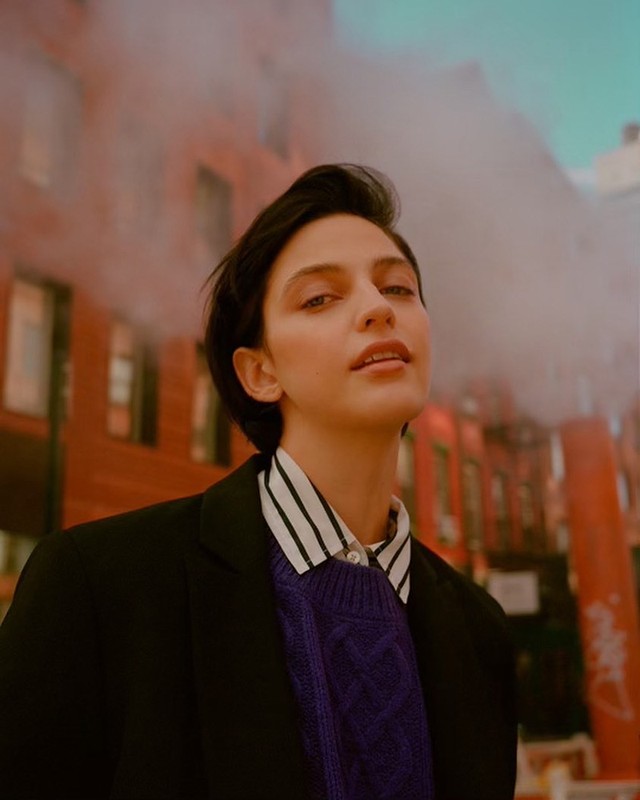 Portrait of a young adult woman with short dark hair in a stylish sweater and jacket, posing against a red brick building.