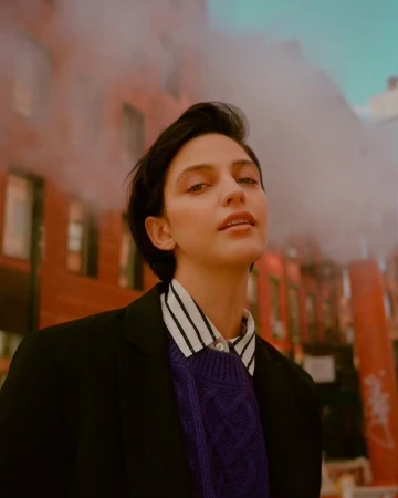 Portrait of a young adult woman with short dark hair in a stylish sweater and jacket, posing against a red brick building.