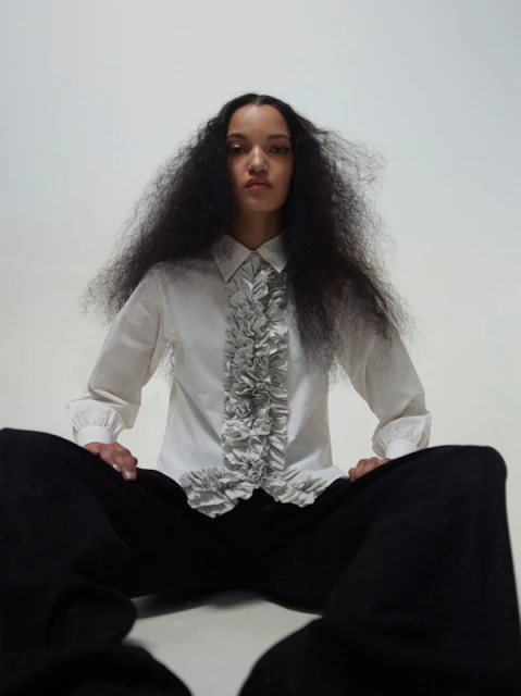 Portrait of a woman with voluminous dark hair posing in a white ruffled shirt and black pants against a light grey studio background.