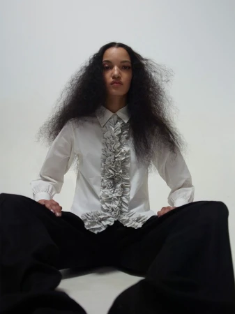 Portrait of a woman with voluminous dark hair posing in a white ruffled shirt and black pants against a light grey studio background.