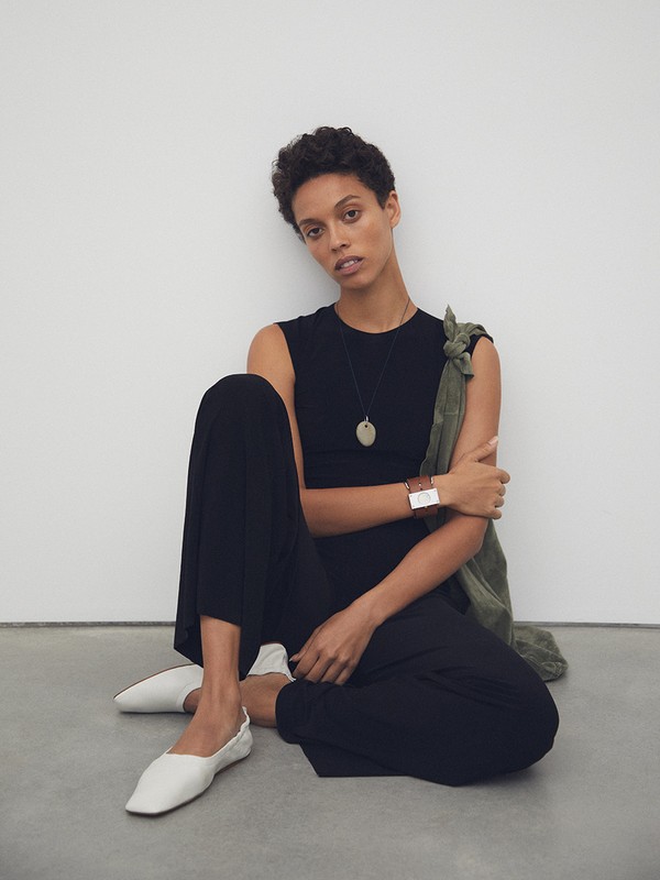 A woman in a black jumpsuit poses while seated on the floor against a white wall, wearing white flats and an oval pendant necklace.