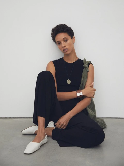 A woman in a black jumpsuit poses while seated on the floor against a white wall, wearing white flats and an oval pendant necklace.