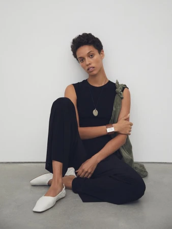 A woman in a black jumpsuit poses while seated on the floor against a white wall, wearing white flats and an oval pendant necklace.