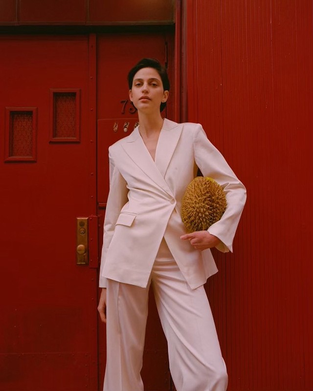 A woman in a white suit stands in front of a red door, holding a spiky, round, yellow object, with a confident expression.