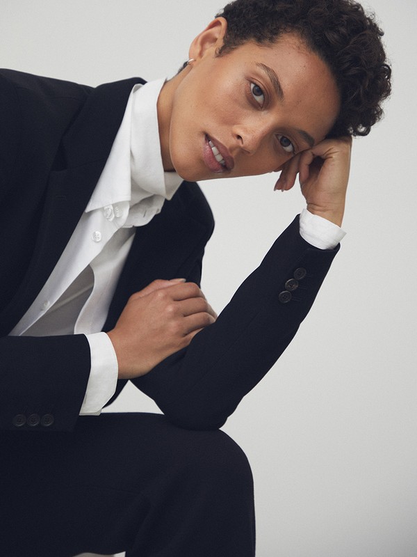 A person with short curly hair wearing a black blazer and white collared shirt poses against a neutral white background.