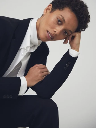 A person with short curly hair wearing a black blazer and white collared shirt poses against a neutral white background.