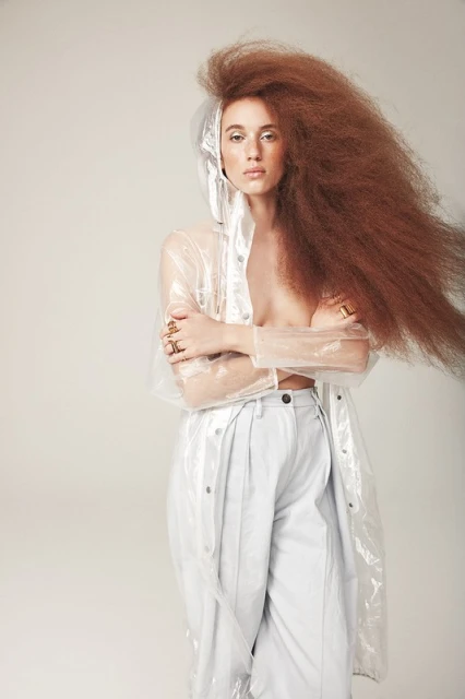 Portrait of a woman with large brown afro hair, wearing a clear raincoat and light colored pants, posing in a photo studio.
