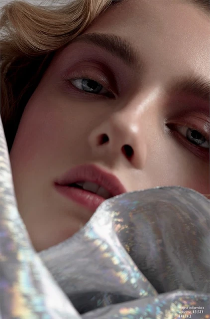 Close-up portrait of a fair-skinned woman with curly blonde hair partially covered in iridescent, shiny fabric against a neutral background.