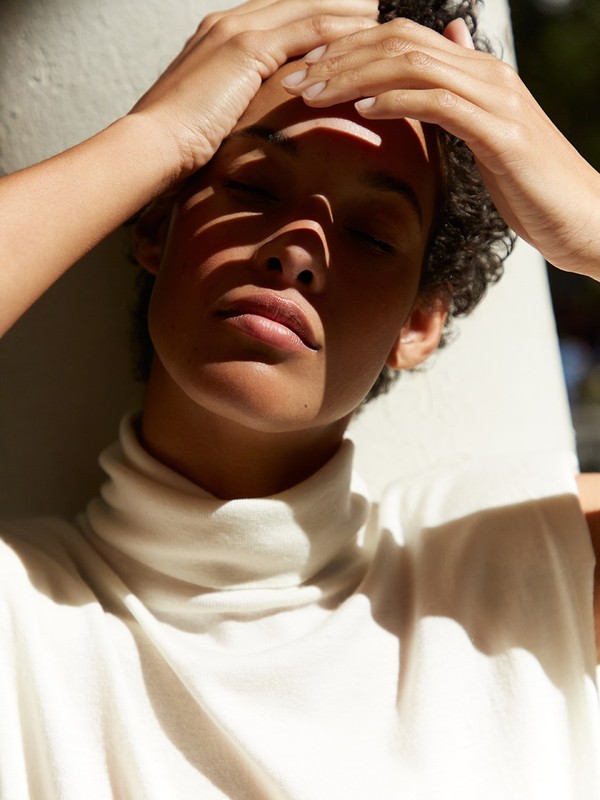 Close-up portrait of an adult woman with curly hair posing in front of a white wall with natural light casting shadows on her face.