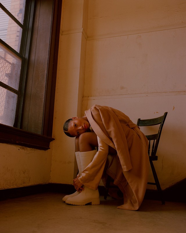 Woman in a beige coat and white boots sits on a chair in an indoor corner, near a window, with muted, warm lighting and fashion styling.