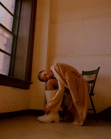Woman in a beige coat and white boots sits on a chair in an indoor corner, near a window, with muted, warm lighting and fashion styling.