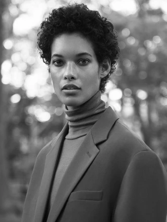 Portrait of a woman with short curly dark hair wearing a turtleneck and blazer, outdoors with bokeh background