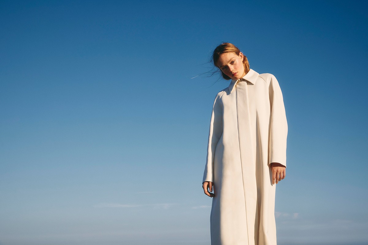 A woman in a cream-colored coat stands in front of a clear blue sky in a fashion shot, looking at the camera at eye-level.