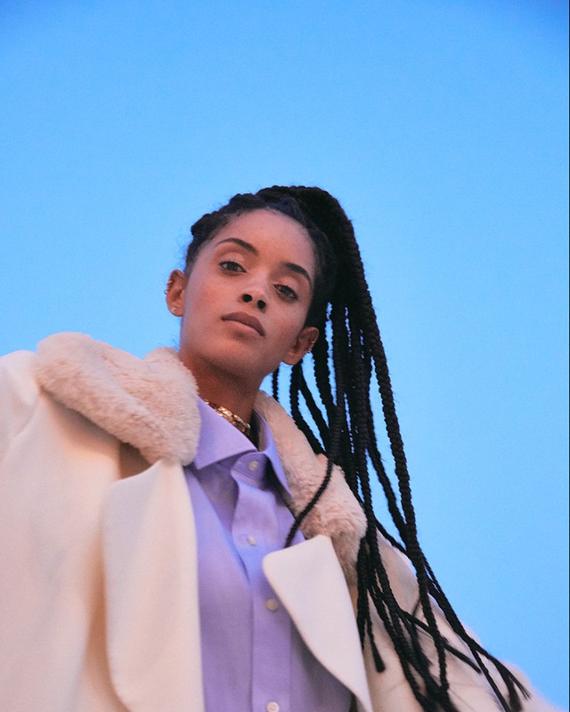 Portrait of a woman in a white coat and lilac shirt looking upward against a blue sky backdrop. Braids, gold necklace, natural light.