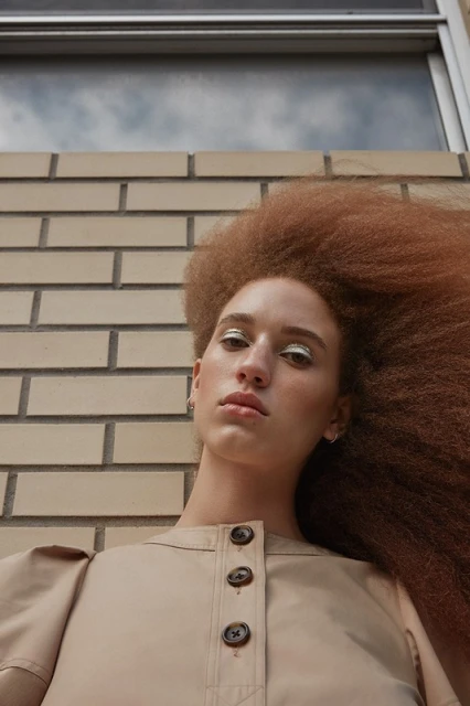 Low-angle shot of a woman with an afro hairstyle wearing a beige collared shirt against a brick wall with a cloudy sky above.