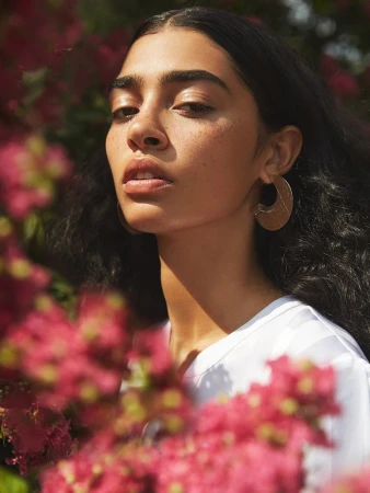 Close-up portrait of a woman with dark hair and hoop earrings framed by pink flowers under natural sunlight