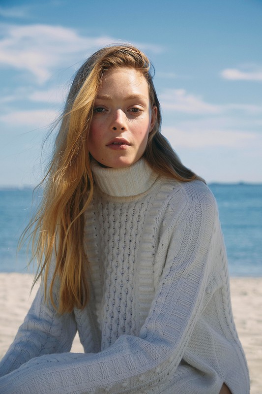 Close-up of a fair-skinned young woman with long blonde hair wearing a white cable-knit turtleneck sweater, posing on a sandy beach.