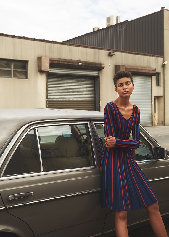 Fashionable woman in striped dress leans against classic car in front of commercial building in daytime urban setting with overcast sky.