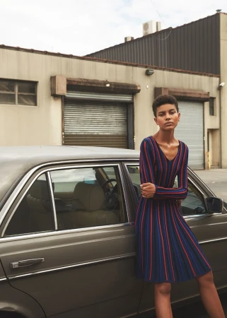 Fashionable woman in striped dress leans against classic car in front of commercial building in daytime urban setting with overcast sky.